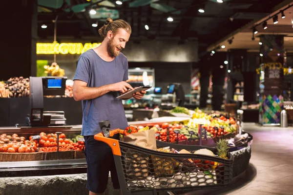 Young smiling man with trolley full of products happily using the tablet for reading shopping list in modern supermarket