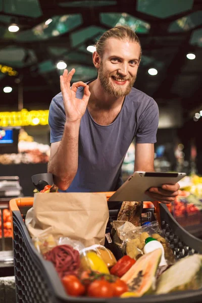 Young smiling man with trolley full of food happily looking in camera showing okay gesture with tablet in hand in modern supermarket