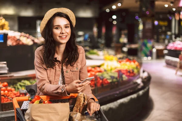 漂亮的微笑的女孩在帽子上靠在车上的所有产品愉快地看着在相机与手机在现代超市的手 — 图库照片