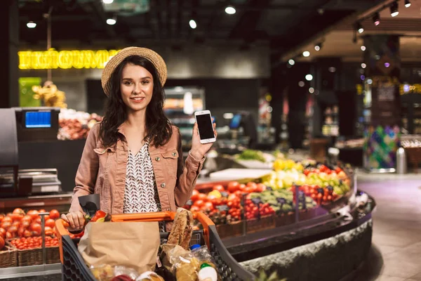 漂亮的微笑的女孩在帽子与手推车充满产品快乐地看着在现代超市中显示新手机的相机 — 图库照片