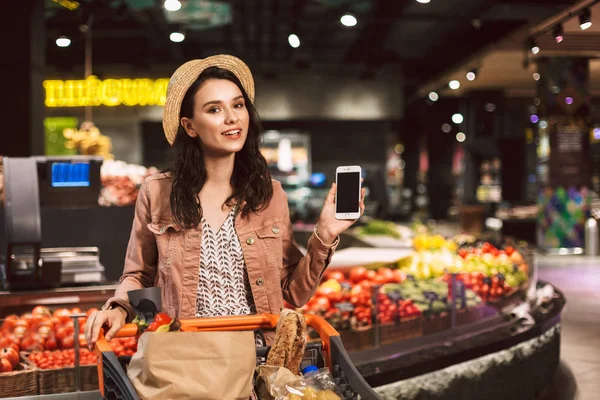 Genç gülümseyen Bayan arabası tam sersem kamera seyir gösteren yeni cep telefonu modern süpermarket ürünleri ile şapkalı