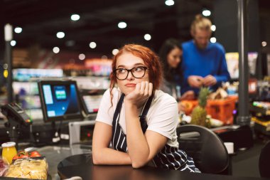 Young boring cashier in eyeglasses and striped apron dreamily looking aside working in modern supermarket with customers on background clipart