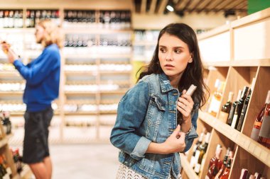 Denim ceket çalışırken genç korkmuş kadın çalmak modern süpermarkette şarap şişe