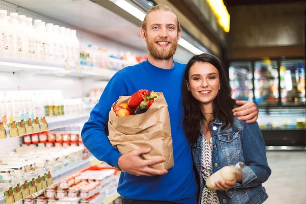 年轻美丽的夫妇与纸杂货袋充满产品愉快地看着在乳品部现代超市的相机 — 图库照片