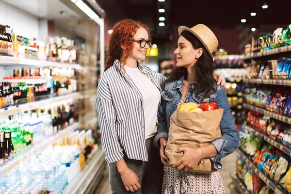 年轻的微笑的女孩与纸杂货袋充满产品愉快地看着彼此在现代超市的花费时间 — 图库照片