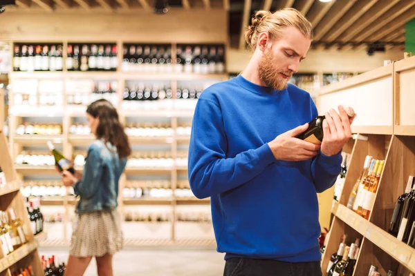 Young Man Dark Blue Sweater Thoughtfully Reading Label Wine Modern — Stock Photo, Image