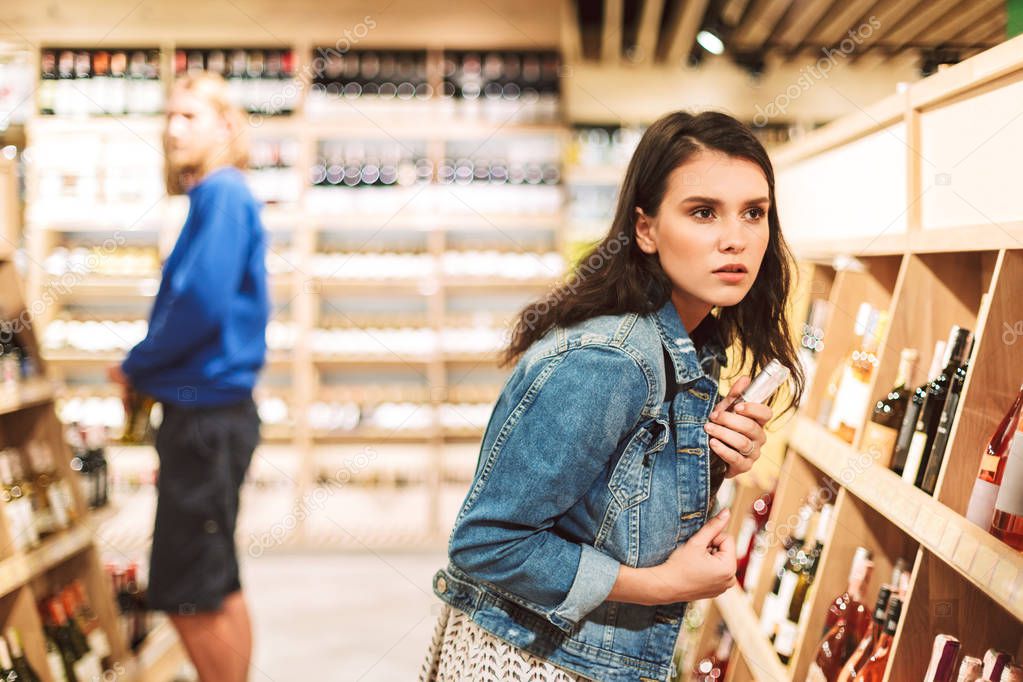 Young serious woman in denim jacket frightenedly looking aside trying steal bottle of wine in modern supermarket