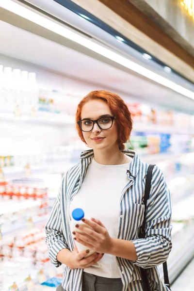 年轻妇女在眼镜和条纹衬衣拿着瓶牛奶在手若有所思地看在现代超级市场的照相机 — 图库照片
