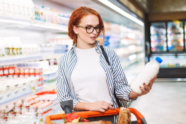 漂亮的女孩在眼镜和条纹衬衫与购物车若有所思地看在现代超市手中的牛奶瓶 — 图库照片