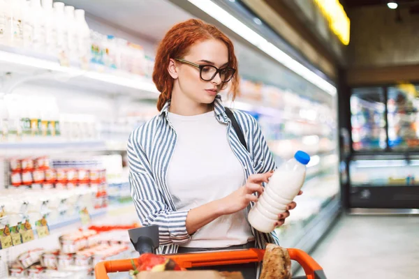 Genç kadın gözlük ve alışveriş sepeti düşünceli şişe süt modern süpermarkette seyir ile çizgili gömlek