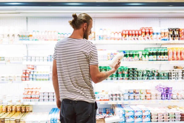 穿条纹 T恤的年轻人若有所思地选择牛奶在现代超市的乳制品部门 — 图库照片