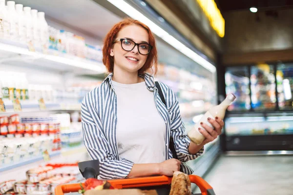 微笑的女孩在眼镜和条纹衬衫与购物车快乐地看着在现代超市手捧着一瓶牛奶的相机 — 图库照片
