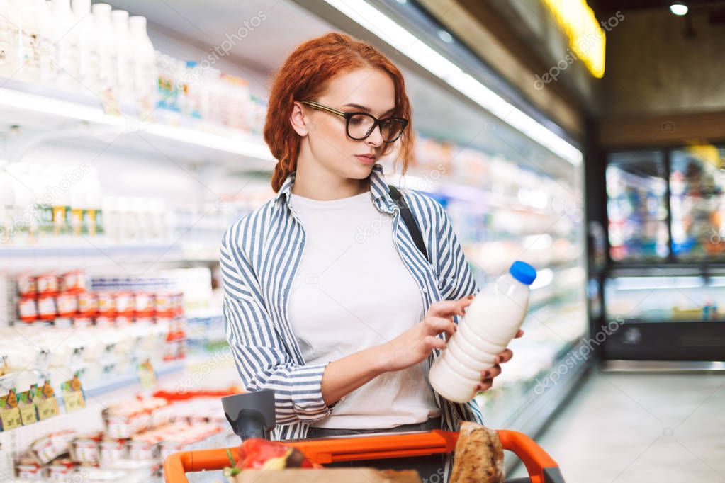 Young woman in eyeglasses and striped shirt with shopping cart thoughtfully looking on bottle of milk in modern supermarket
