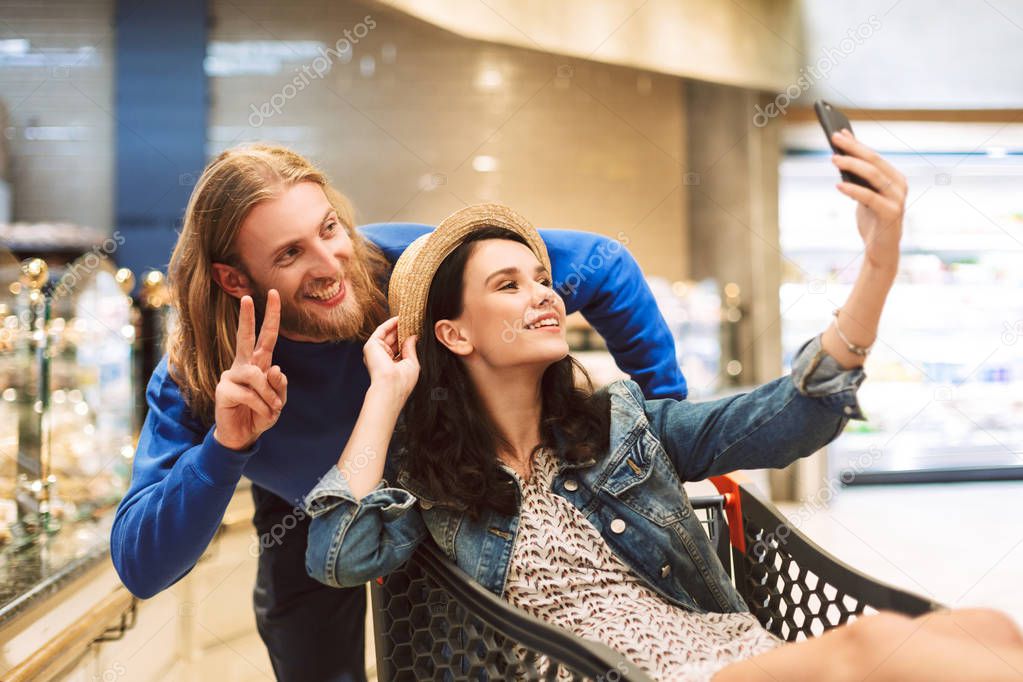 Young cheerful guy with pretty girl inside shopping trolley taking cute photos together on cellphone in modern supermarket