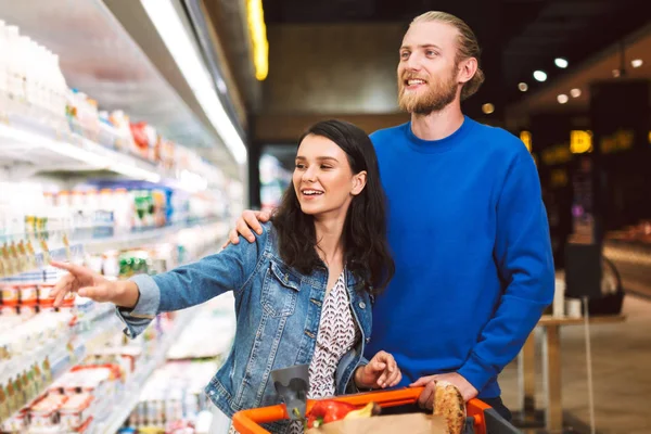 Young joyful couple with trolley full of products happily choosing dairy products in modern supermarket
