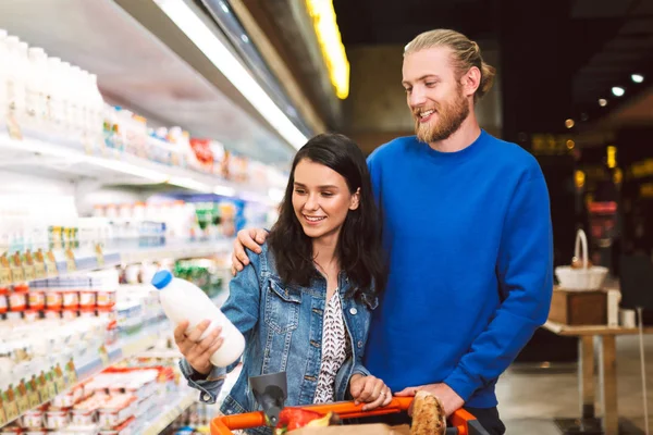 年轻的微笑夫妇与手推车装满产品在现代超市中快乐地选择牛奶 — 图库照片