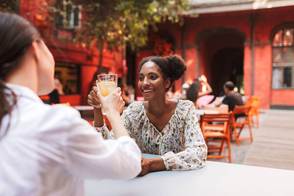 Pretty smiling african girl in dress drinking cocktails with friend joyfully spending time together in cozy courtyard of cafe