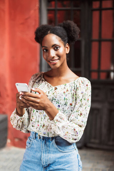 Beautiful smiling african girl in blouse and denim skirt holding cellphone in hands happily looking in camera outdoor