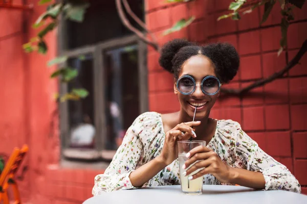 Young african woman in blouse and sunglasses happily looking in camera drinking cocktail in cozy courtyard of cafe