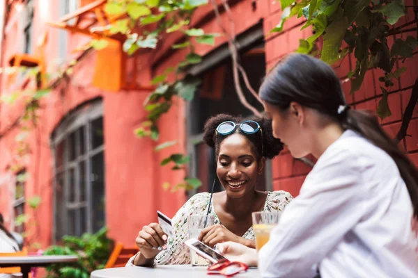 Pretty smiling girls using cellphone and credit card happily spending time together in cozy courtyard of cafe