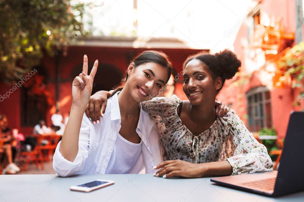 Pretty smiling girls friendly hugging each other happily showing two fingers in camera with laptop and cellphone on table in cozy courtyard of cafe 
