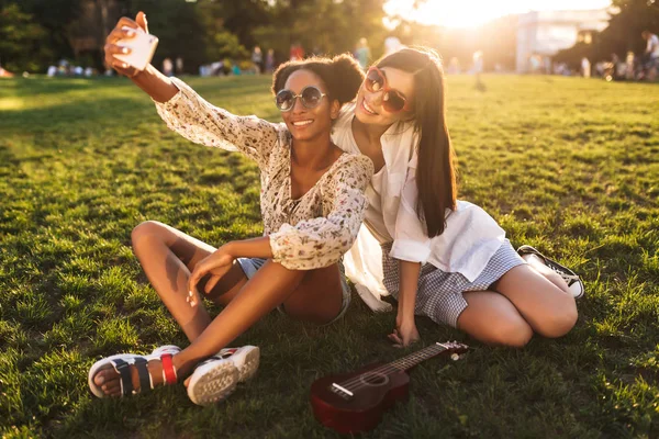 美丽的微笑的女孩坐在草地上快乐地拍照手机花时间一起在城市公园 — 图库照片