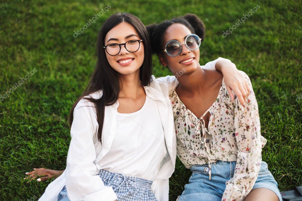 Two cheerful girls friendly hugging each other happily looking in camera spending time together on grass in park