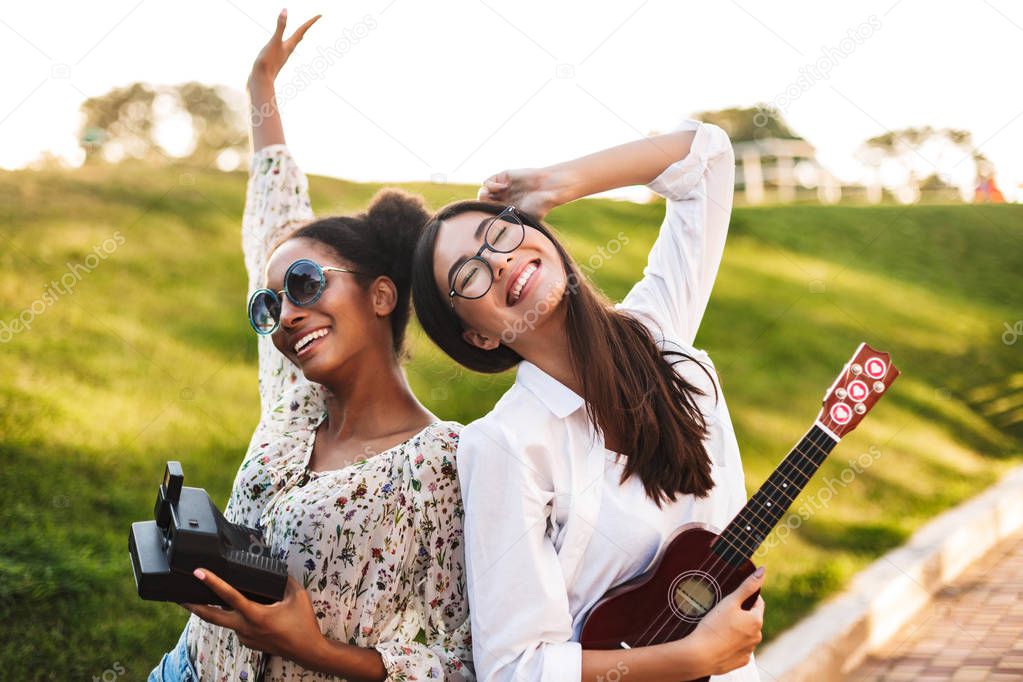 Cheerful girls with little guitar and polaroid camera in hands joyfully spending time together in park