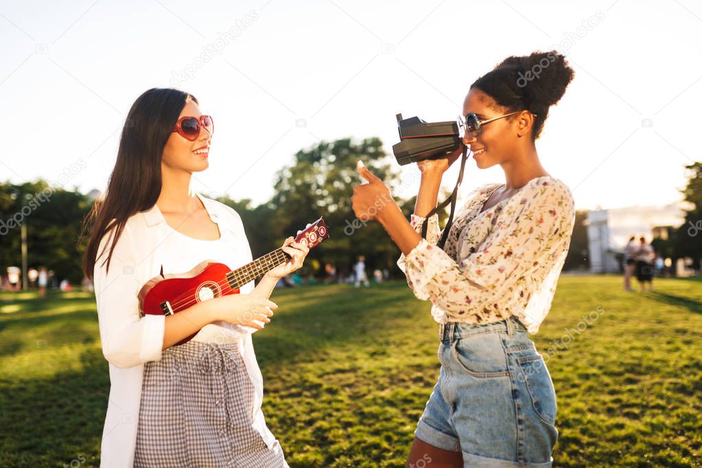 Beautiful girl taking photos on polaroid camera while pretty girl in sunglasses posing with little guitar in park
