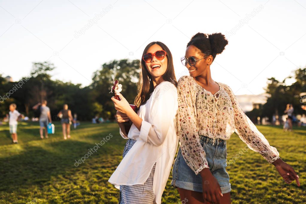Beautiful girls in sunglasses happily playing on little guitar and dancing spending time together in park