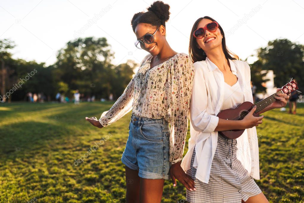 Joyful girls in sunglasses happily playing on little guitar and dancing spending time together in city park