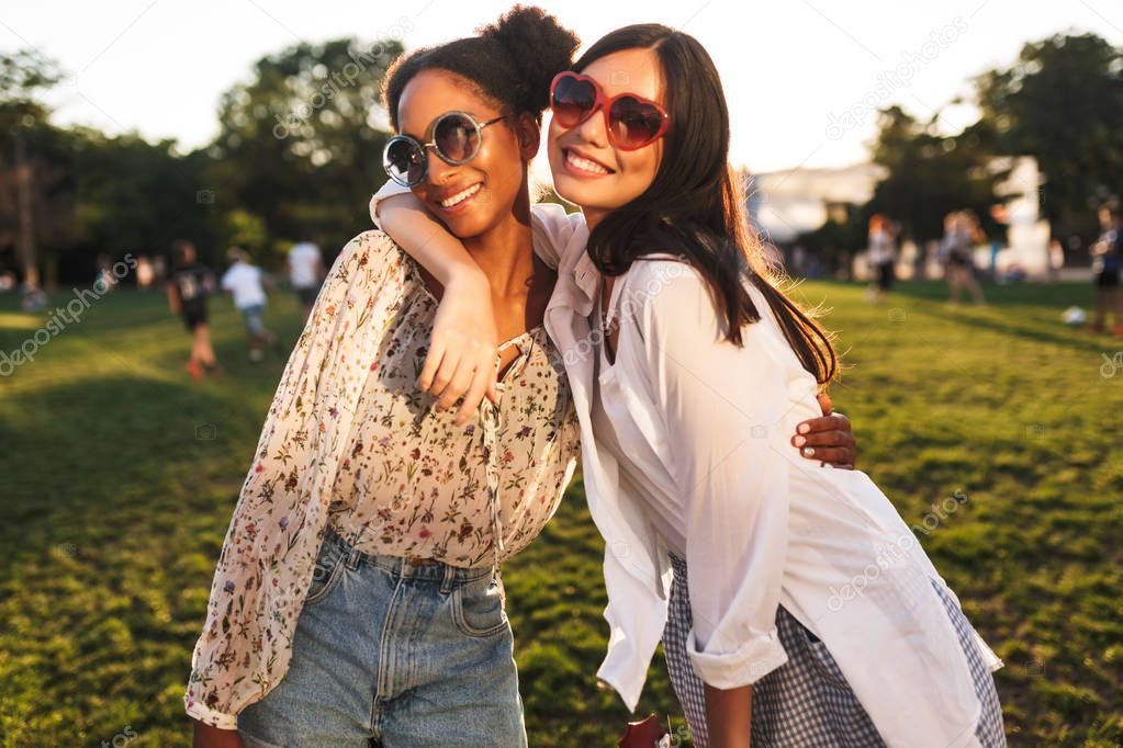 Two pretty girls in sunglasses friendly hugging each other happily looking in camera spending time together in city park