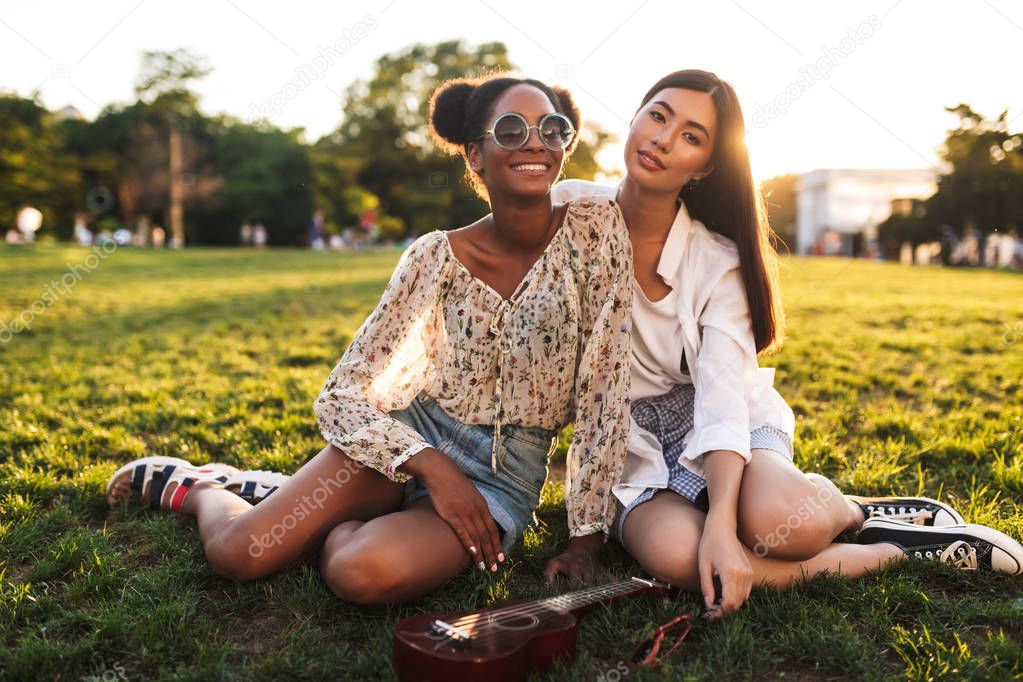 Pretty ladies sitting on grass dreamily looking in camera spending time together in city park