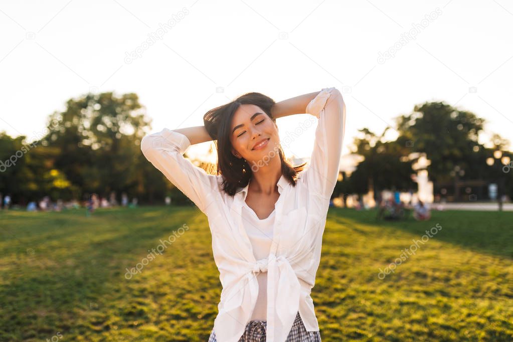 Pretty smiling asian girl in white shirt dreamily closing eyes happily spending time in park