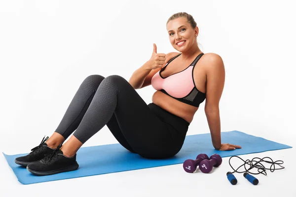 Pretty smiling girl with excess weight in sporty top and leggings sitting on yoga mat happily showing thumb up gesture looking in camera over white background. Plus size model