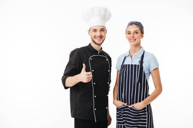 Siyah üniforma ve beyaz şapka ve güzel kadın genç gülümseyen adam şef çizgili önlük yemek ve mutlu bir şekilde fotoğraf makinesindeki beyaz arka plan üzerinde başparmak gösterilmesini seyir kap