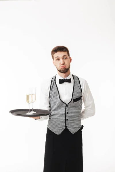 Young waiter in uniform holding tray with glasses of champagne tiredly looking in camera over white background