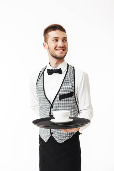 Молодой радостный официант в форме держит поднос с чашкой кофе в руке счастливо глядя в сторону на белом фоне
