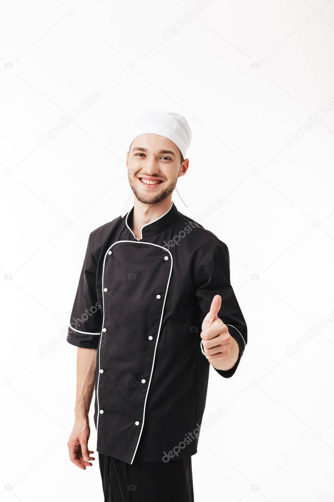 Young smiling man chef in black uniform joyfully looking in camera showing thumb up gesture over white background