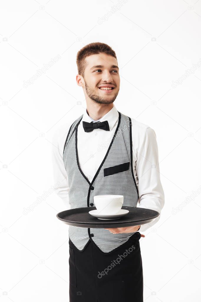 Young joyful waiter in uniform holding tray with cup of coffee in hand happily looking aside over white background