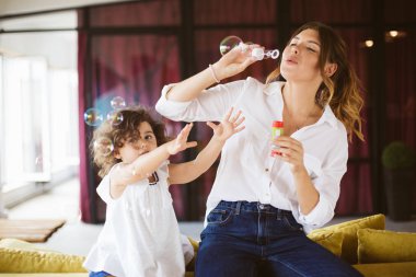 Mutlu birlikte vakit kabarcıklar evde üfleme elbiseli küçük şirin kızı ile beyaz gömlekli güzel genç anne 