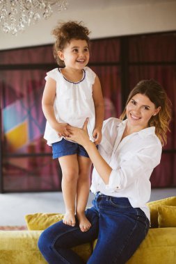 Mutlu kamera seyir kot pantolon ve beyaz gömlek gülümseyen genç anne küçük neşeli kızı evde dizlerinin üzerinde tutarak.