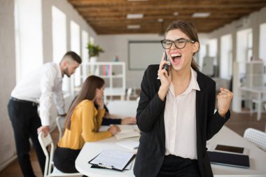 Genç iş kadını gözlük ve duygusal ofis cep telefonu arka plan meslektaşları ile konuşurken çığlık gömlek