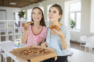 Mutlu bir şekilde vakit modern beyaz ofis ile birlikte arka plan üzerinde büyük lezzetli pizza yemek masasında oturan iki neşeli kız