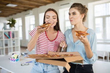Sevinçle modern ofiste birlikte vakit büyük pizza yemek masasında oturan iki güzel kızlar
