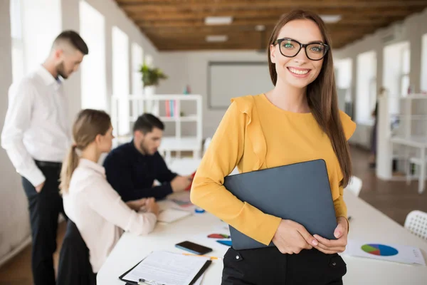Genç gülümseyen iş kadını mutlu kamera tutarak klasörü Office elinde meslektaşları ile arka plan üzerinde seyir gözlük