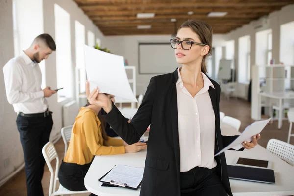 年轻的商业妇女在眼镜和衬衫若有所思地看文件与同事在办公室工作背景 — 图库照片