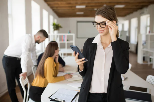 年轻微笑的商业妇女在眼镜和衬衫愉快地使用手机花时间在办公室与同事的背景 — 图库照片
