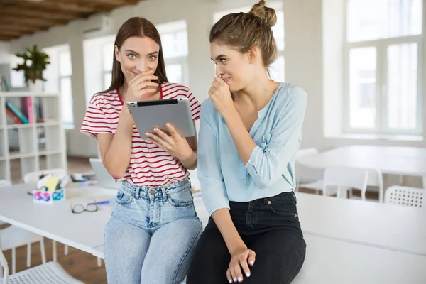 Smiling girls sitting on desk with tablet happily gossiping together with modern empty office on background