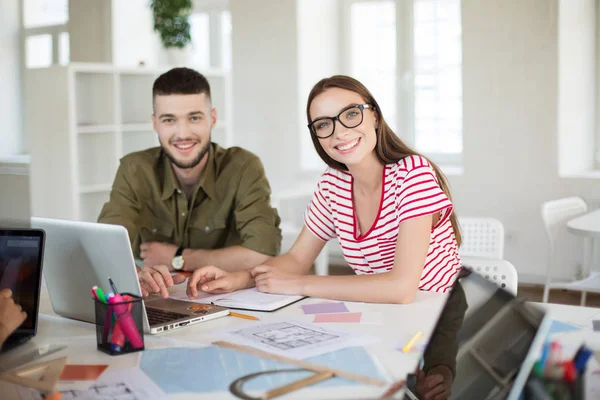 Young man and woman in striped t-shirt and eyeglasses happily looking in camera working together. Group of cool guys with laptop spending time in modern office